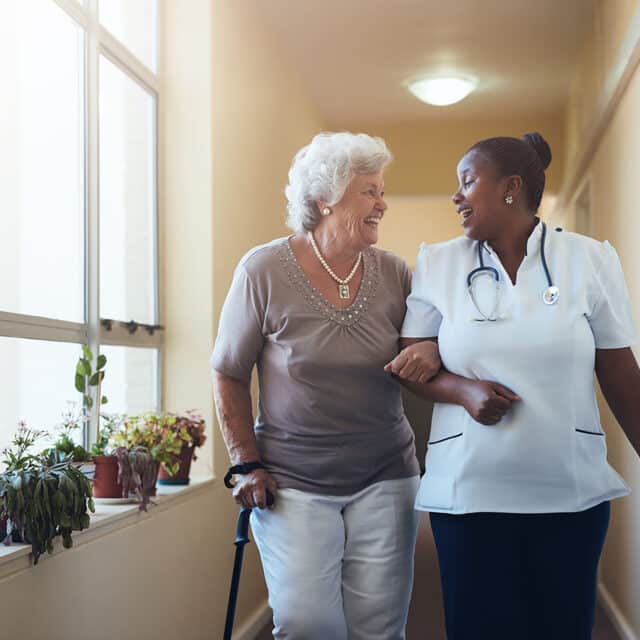 A nurse and an older woman strategically navigate a hallway, showcasing global innovation in patient care.
