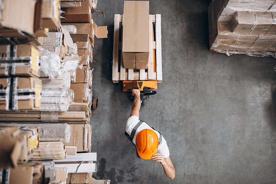 A company worker in an orange hard hat is strategically operating a forklift in a warehouse.