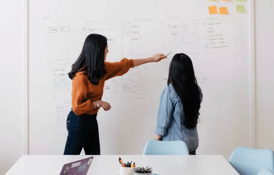 Two women pointing at a whiteboard in a tech office.