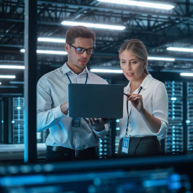 Two tech consultants discussing a laptop in a server room of a company.