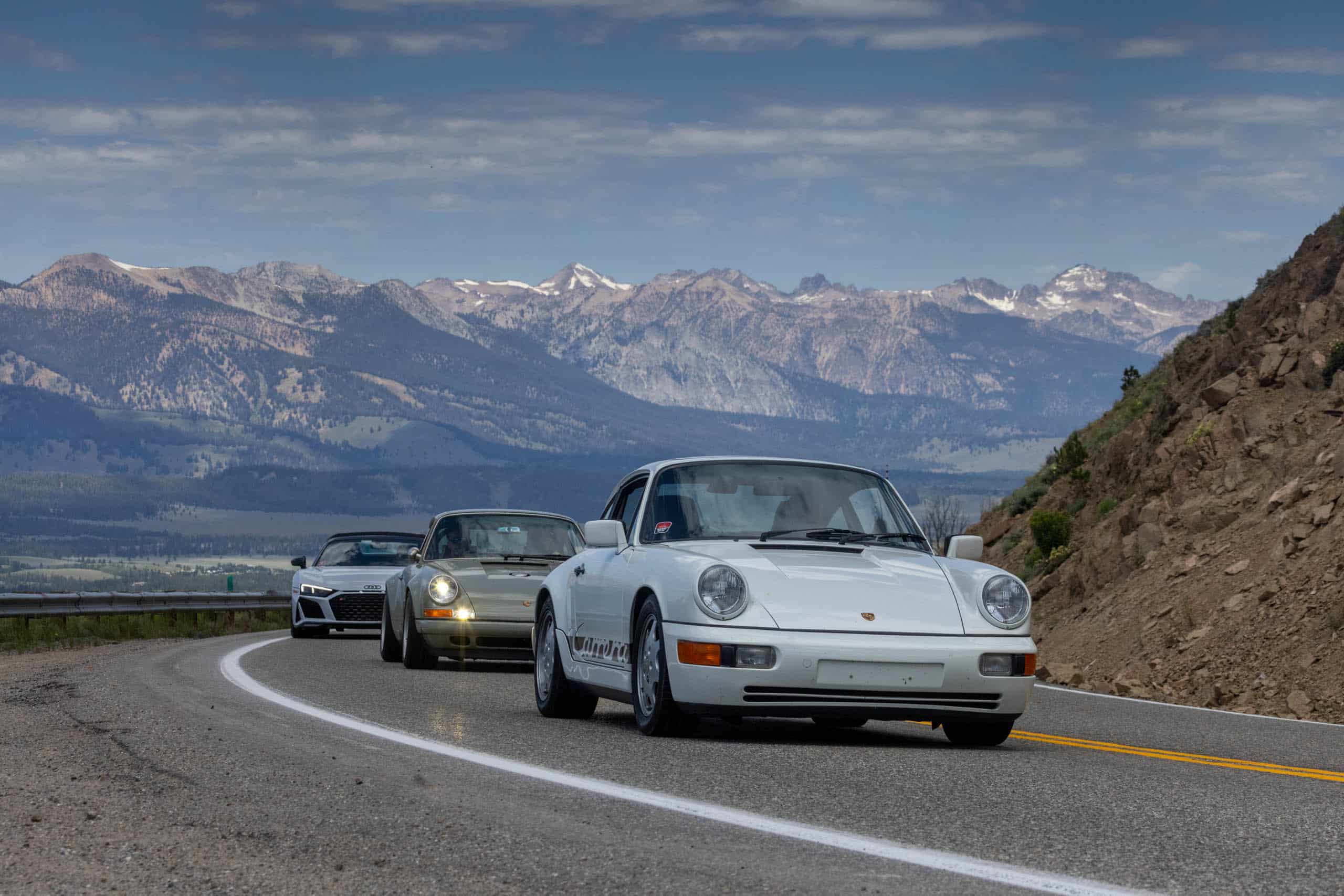 A white porsche 911 driving down a road surrounded by majestic mountains.