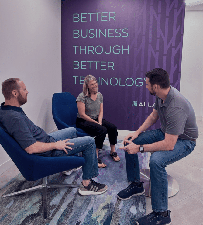 Three people sitting in chairs in an office on the careers page, with a sign saying better business better technology.